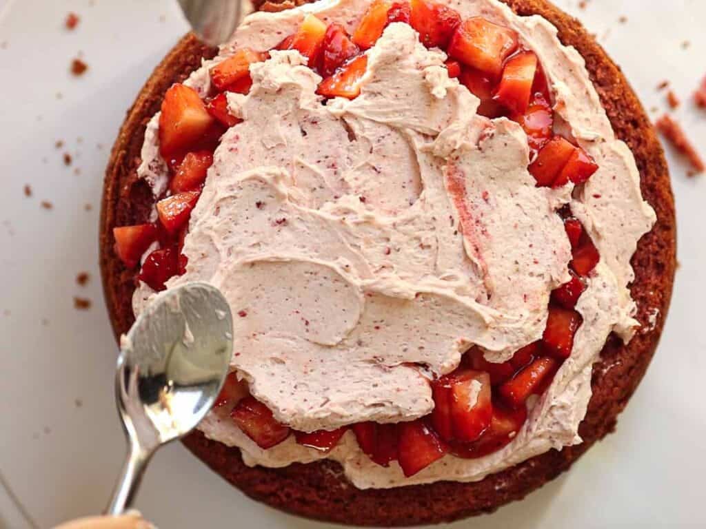 Overhead view of a vegan strawberry cake being frosted with whipped cream and topped with chopped strawberries. Two spoons are spreading the cream over the fruit on the round cake layer. Crumbs are visible around the cake on a white surface.